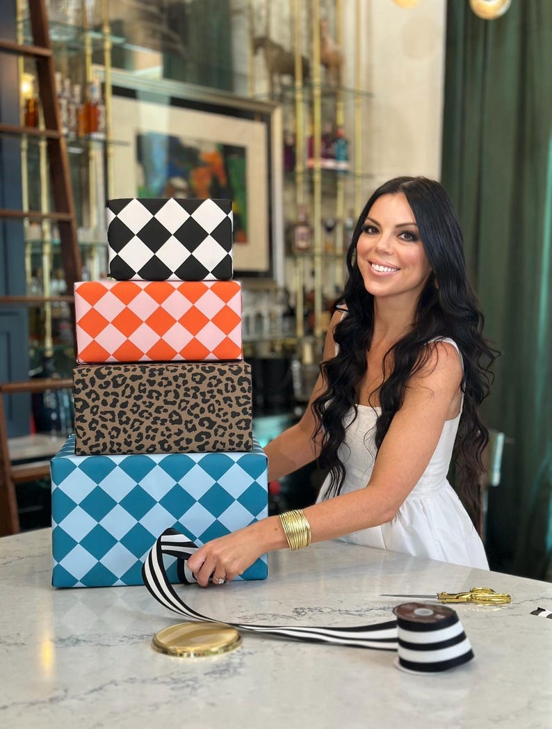 May include: A stack of four wrapped gift boxes with various patterns: black and white diamonds, orange and pink diamonds, leopard print, and blue diamonds. A person is holding a black and white striped ribbon, with gold scissors and a spool of ribbon nearby.