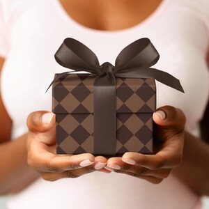 Op de afbeelding: Een persoon houdt een bruin en zwart cadeaudoosje vast met een zwart satijnen lint.