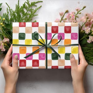 May include: A white gift box with a colorful checkered pattern. The box is wrapped with a green ribbon and tied in a bow. The box is surrounded by green leaves and pink flowers.