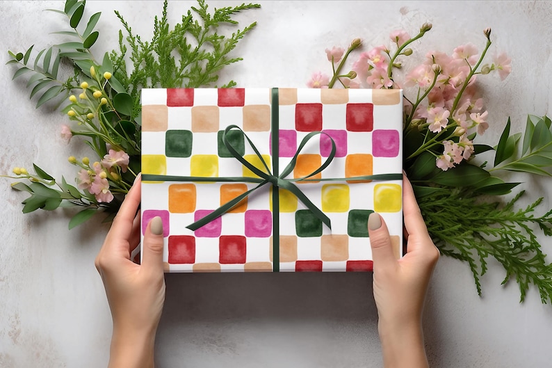 May include: A white gift box with a colorful checkered pattern. The box is wrapped with a green ribbon and tied in a bow. The box is surrounded by green leaves and pink flowers.