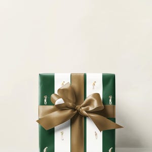 May include: Wrapped gift box with green and white striped paper featuring illustrations of golfers. A wide gold ribbon and bow are tied around the box. The background is a solid off-white color.