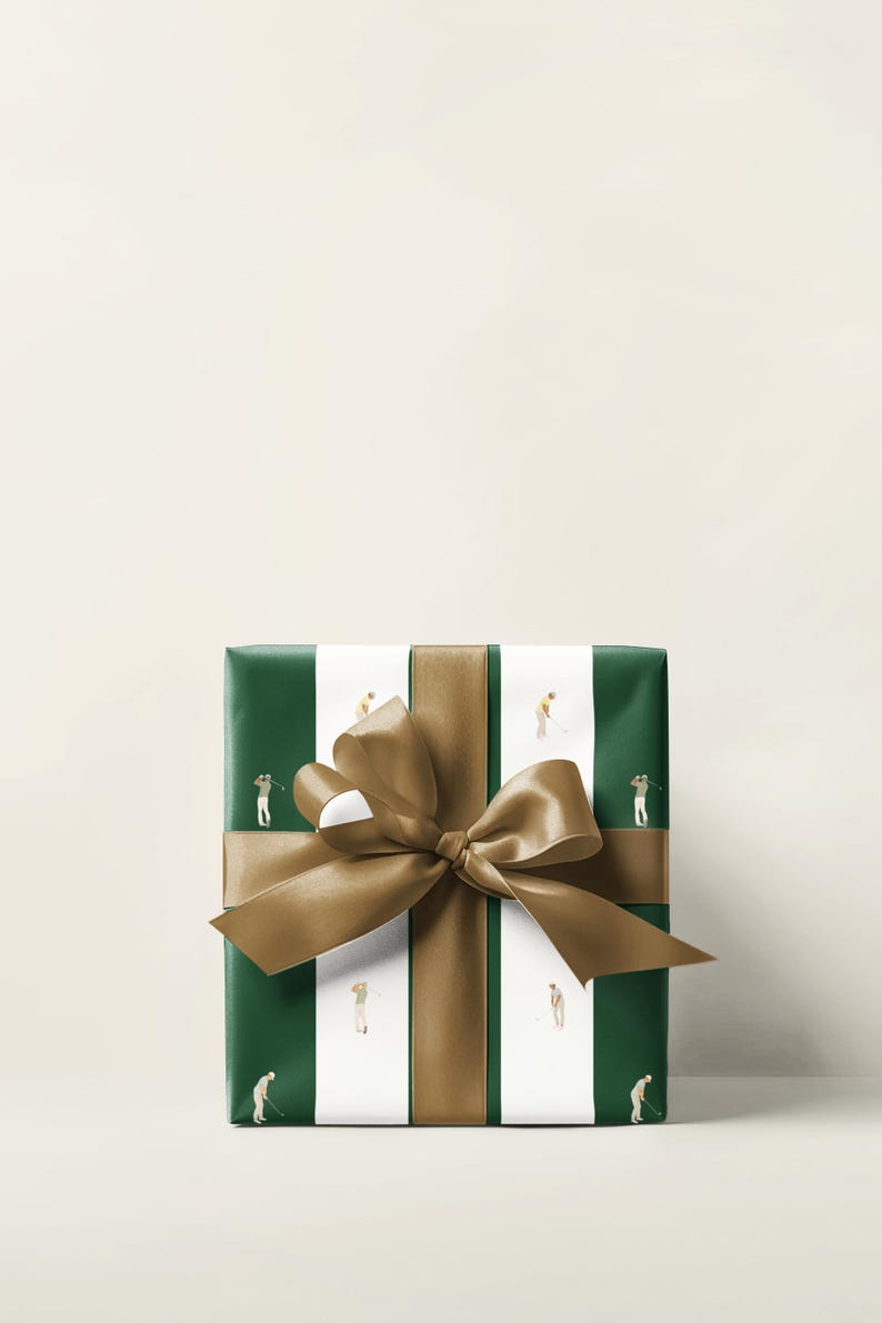 May include: Wrapped gift box with green and white striped paper featuring illustrations of golfers. A wide gold ribbon and bow are tied around the box. The background is a solid off-white color.