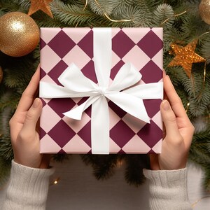K&ouml;nnte beinhalten: Eine pinkfarbene und burgunderrote Geschenkbox mit einem wei&szlig;en Band und Schleife. Die Box ist von einem Weihnachtsbaum mit goldenen Ornamenten und Sternen umgeben.
