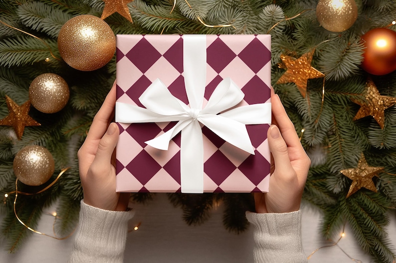 K&ouml;nnte beinhalten: Eine pinkfarbene und burgunderrote Geschenkbox mit einem wei&szlig;en Band und Schleife. Die Box ist von einem Weihnachtsbaum mit goldenen Ornamenten und Sternen umgeben.