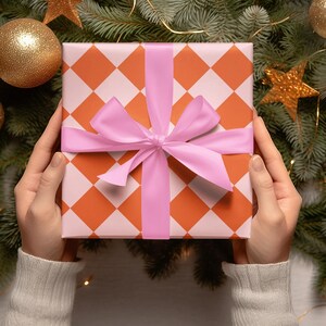 May include: A gift box wrapped in orange and white checkered paper with a pink ribbon. The box is surrounded by a Christmas tree with gold ornaments and lights.