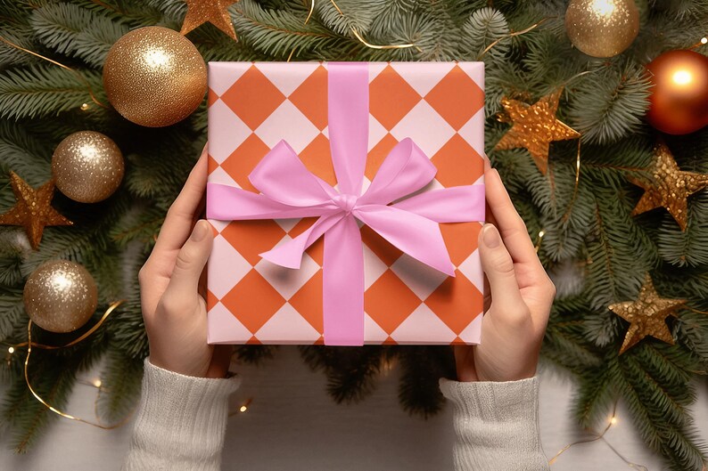 May include: A gift box wrapped in orange and white checkered paper with a pink ribbon. The box is surrounded by a Christmas tree with gold ornaments and lights.