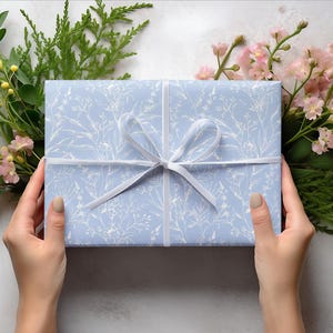 Puede incluir: Una caja de regalo azul claro con una cinta blanca y un patr&oacute;n floral blanco. La caja est&aacute; siendo sostenida por dos manos.