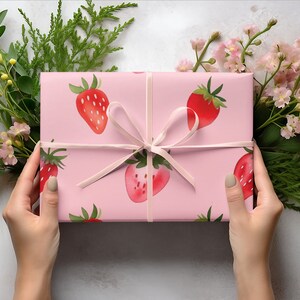 May include: A pink gift box with a white ribbon and a strawberry pattern. The box is wrapped in pink paper with a white ribbon tied in a bow. The paper has a pattern of strawberries on it.