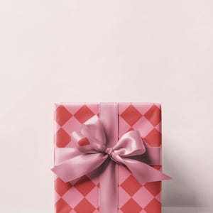 May include: A wrapped gift box with a pink and red diamond pattern. The box is tied with a wide pink satin ribbon and a large bow. The background is a solid light pink.