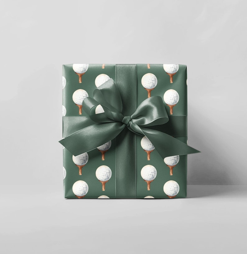K&ouml;nnte beinhalten: Ein verpacktes Geschenk mit gr&uuml;nem Geschenkpapier, das mit wei&szlig;en Golfb&auml;llen auf Abschl&auml;gen gemustert ist. Ein passendes gr&uuml;nes Satinband ist oben zu einer Schleife gebunden. Das Geschenk ist quadratisch.