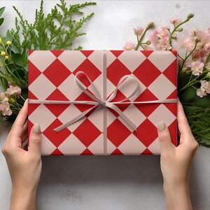 K&ouml;nnte beinhalten: Ein Geschenk, verpackt mit einem rot-wei&szlig; karierten Muster, gebunden mit einem grauen Band. Das Geschenk ist von Gr&uuml;n und rosa Bl&uuml;ten umgeben, gehalten von zwei H&auml;nden.
