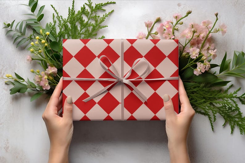 K&ouml;nnte beinhalten: Ein Geschenk, verpackt mit einem rot-wei&szlig; karierten Muster, gebunden mit einem grauen Band. Das Geschenk ist von Gr&uuml;n und rosa Bl&uuml;ten umgeben, gehalten von zwei H&auml;nden.