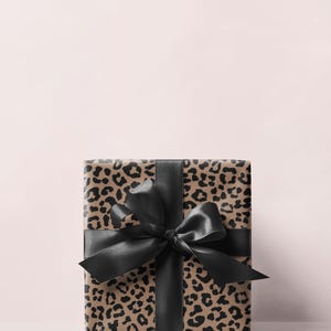 May include: A wrapped gift box featuring a leopard print design in shades of brown and black. The box is tied with a wide, black satin ribbon and bow. The background is a soft pink.