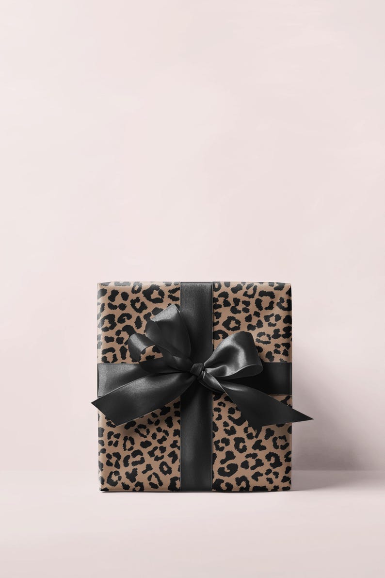 May include: A wrapped gift box featuring a leopard print design in shades of brown and black. The box is tied with a wide, black satin ribbon and bow. The background is a soft pink.