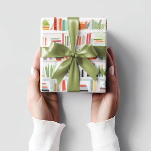 May include: A wrapped gift with a green satin ribbon and bow. The wrapping paper features a repeating pattern of bookshelves with books and plants in various colors. The gift is held by two hands against a light gray background.