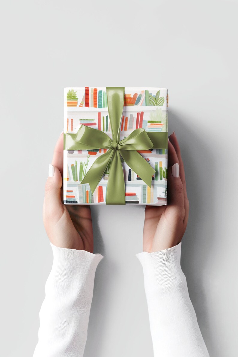 May include: A wrapped gift with a green satin ribbon and bow. The wrapping paper features a repeating pattern of bookshelves with books and plants in various colors. The gift is held by two hands against a light gray background.