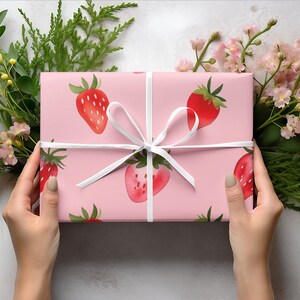 May include: A pink gift box with a white ribbon and a strawberry pattern. The box is being held by two hands.
