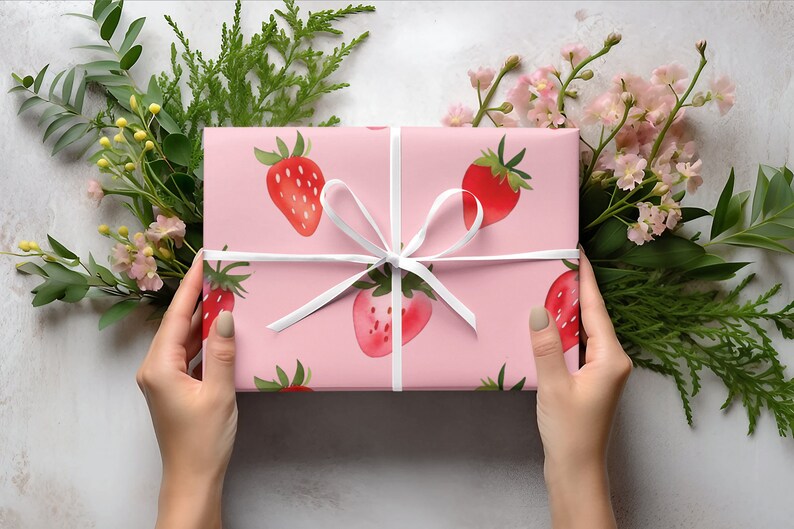 May include: A pink gift box with a white ribbon and a strawberry pattern. The box is being held by two hands.