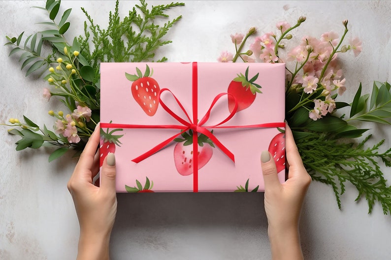 May include: A pink gift box with a red ribbon and a strawberry pattern. The box is wrapped in pink paper with red strawberries printed on it.