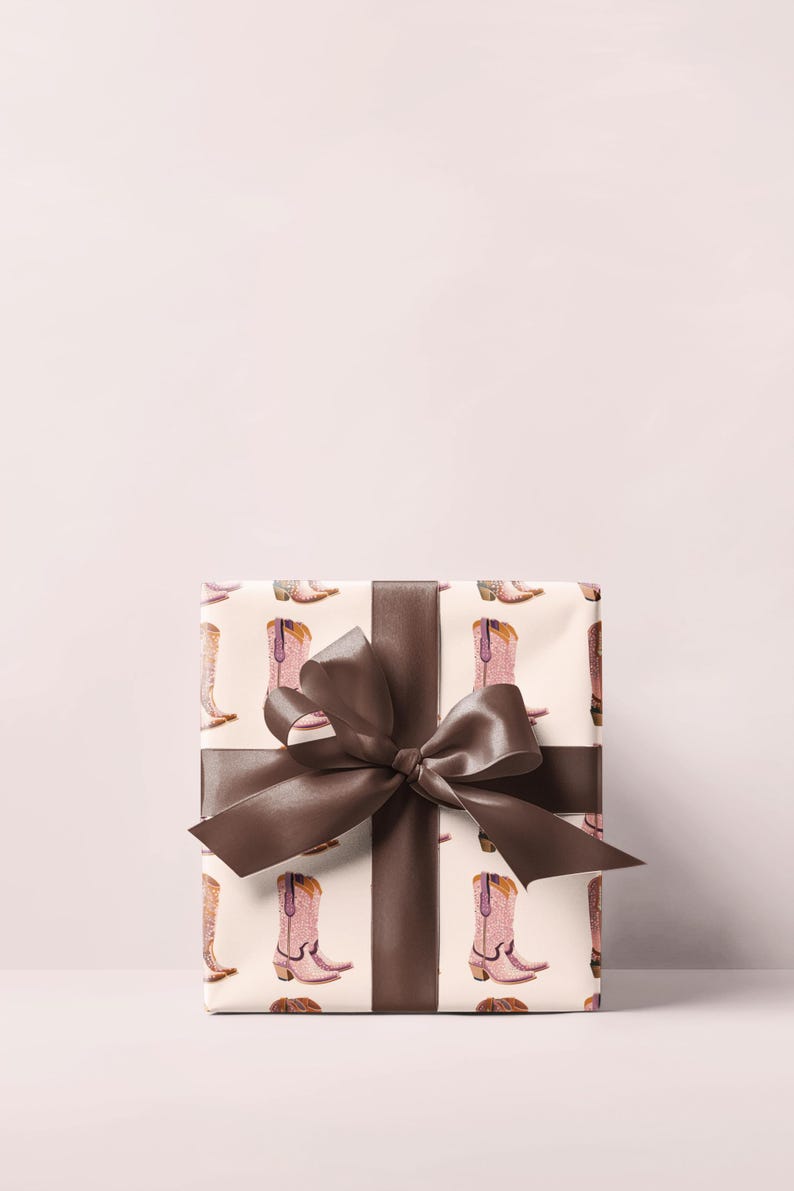 Pu&ograve; includere: Una scatola regalo bianca con un nastro e un fiocco marroni. La scatola &egrave; avvolta in carta con un motivo di stivali da cowboy rosa.
