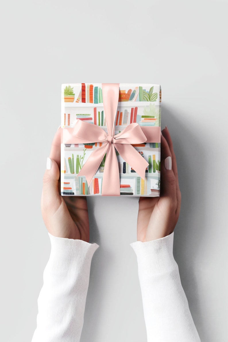 May include: A wrapped gift with a pink satin ribbon. The wrapping paper features a repeating pattern of colorful bookshelves with books and plants. The gift is held by two hands, with white sleeves and a light gray background.
