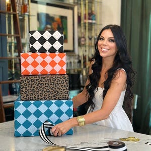 May include: A stack of four gift boxes with patterned wrapping paper. The patterns include black and white diamonds, orange and pink diamonds, leopard print, and blue diamonds. A person is wrapping a gift with black and white striped ribbon.
