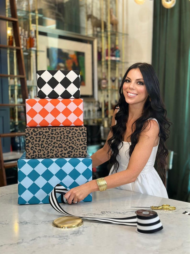 May include: A stack of four gift boxes with patterned wrapping paper. The patterns include black and white diamonds, orange and pink diamonds, leopard print, and blue diamonds. A person is wrapping a gift with black and white striped ribbon.