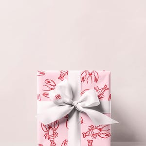 May include: A wrapped gift box with pink wrapping paper featuring a red lobster pattern. The gift is tied with a wide white satin ribbon and bow. The background is a solid light pink.