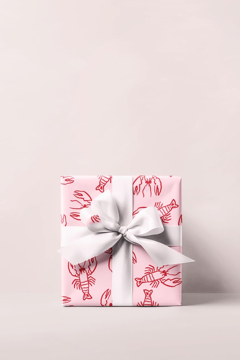 May include: A wrapped gift box with pink wrapping paper featuring a red lobster pattern. The gift is tied with a wide white satin ribbon and bow. The background is a solid light pink.
