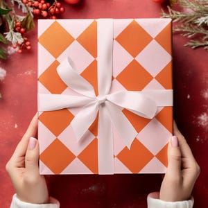 May include: A gift box with a pink and orange checkered pattern is wrapped with a white satin ribbon. The box is held by hands wearing white sweaters.