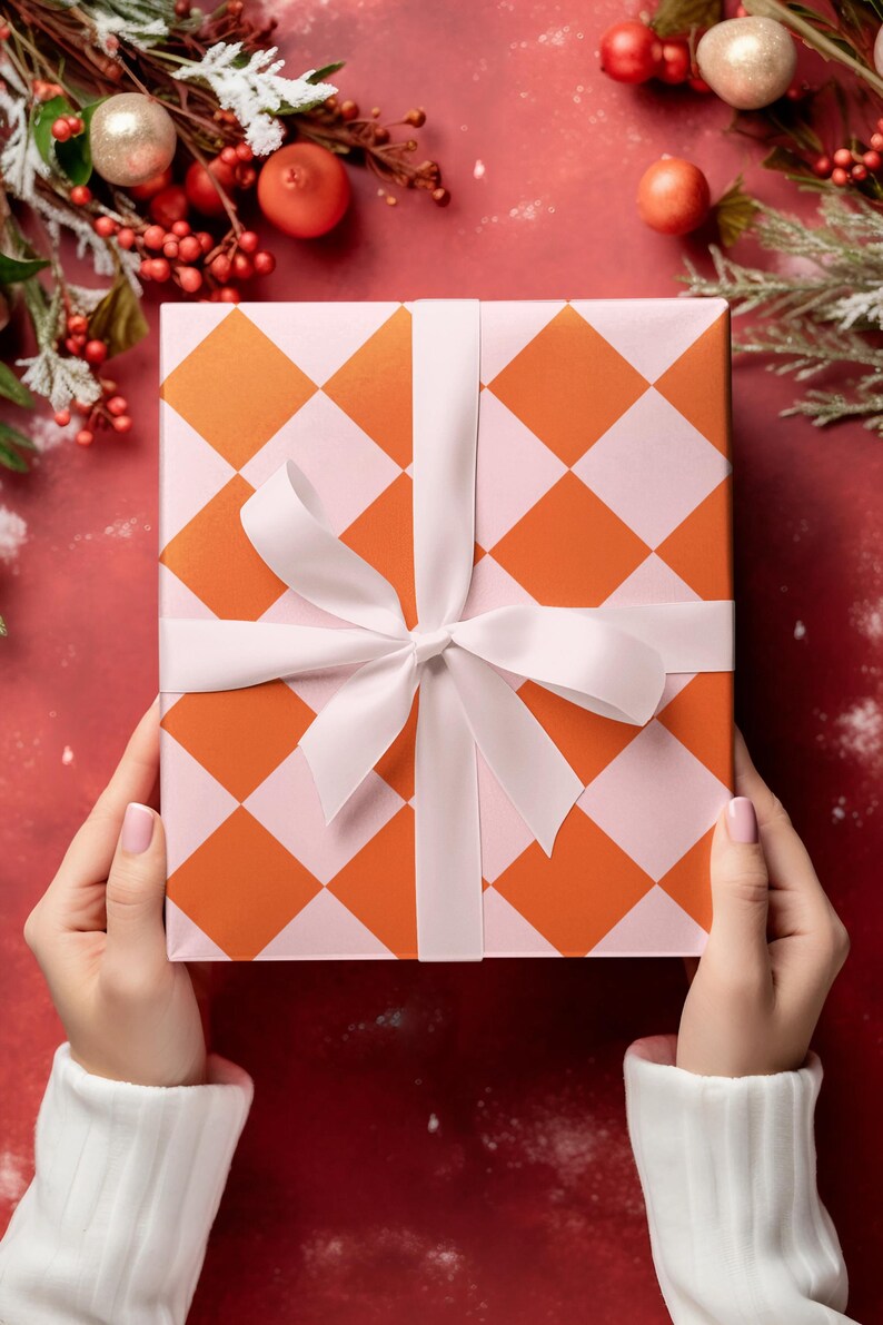 May include: A gift box with a pink and orange checkered pattern is wrapped with a white satin ribbon. The box is held by hands wearing white sweaters.
