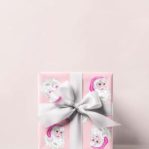 Puede incluir: Una caja de regalo envuelta con papel rosa con un patr&oacute;n repetido de caras de Pap&aacute; Noel. La caja est&aacute; atada con una cinta y un lazo de sat&eacute;n blanco. El fondo es de color rosa claro.