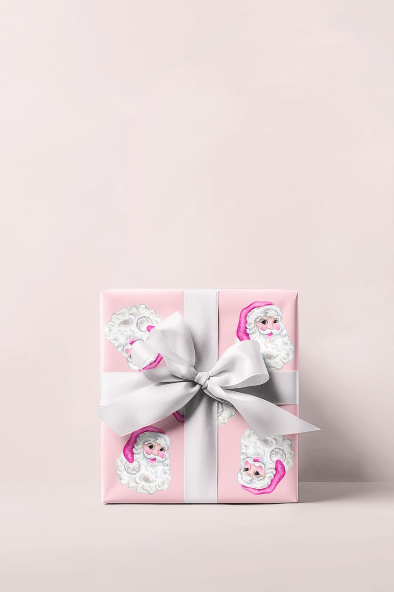 Puede incluir: Una caja de regalo envuelta con papel rosa con un patr&oacute;n repetido de caras de Pap&aacute; Noel. La caja est&aacute; atada con una cinta y un lazo de sat&eacute;n blanco. El fondo es de color rosa claro.