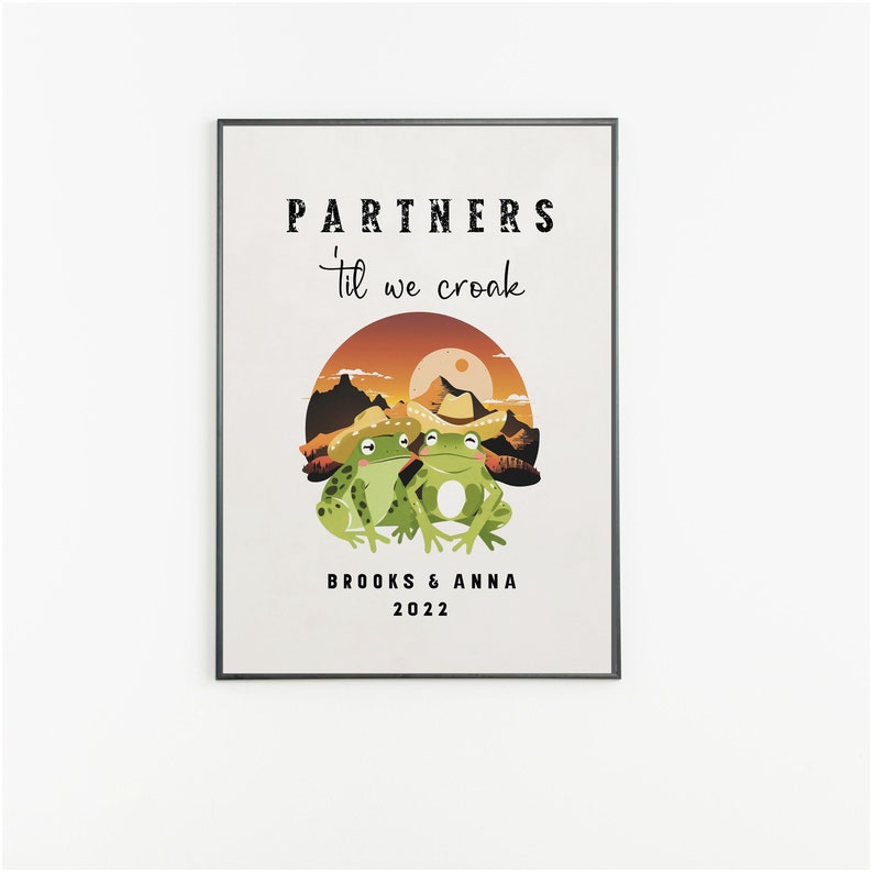 May include: A framed art print featuring two green frogs wearing cowboy hats, set against a sunset landscape. The text reads "PARTNERS 'til we croak", with the names "BROOKS & ANNA" and the year "2022" below the image. The artwork is displayed on a white wall.
