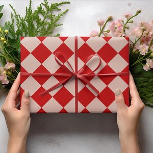 K&ouml;nnte beinhalten: Ein Geschenk, verpackt mit einem rot-wei&szlig; karierten Muster, gebunden mit einem roten Band. Das Geschenk ist von Gr&uuml;n und rosa Blumen umgeben. Das Geschenk wird von zwei H&auml;nden gehalten.
