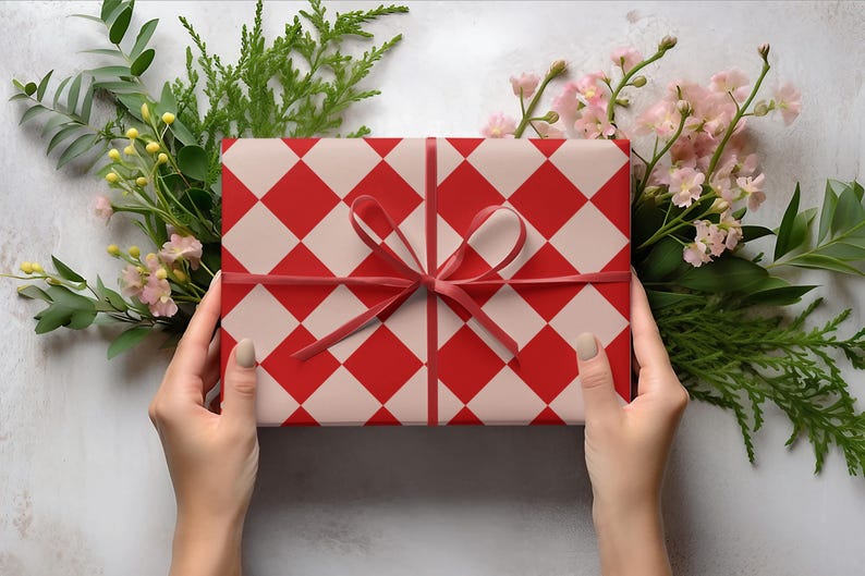 K&ouml;nnte beinhalten: Ein Geschenk, verpackt mit einem rot-wei&szlig; karierten Muster, gebunden mit einem roten Band. Das Geschenk ist von Gr&uuml;n und rosa Blumen umgeben. Das Geschenk wird von zwei H&auml;nden gehalten.