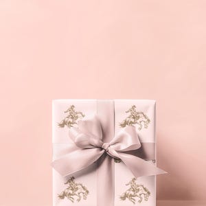 May include: A wrapped gift box with a light pink background. The gift wrap is white with a repeating gold illustration of a cowboy riding a horse. A light pink ribbon and bow are tied around the box.