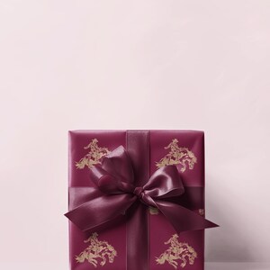 May include: A burgundy gift box with a matching ribbon and bow. The wrapping paper has a repeating gold illustration of a cowboy on a horse. The gift is set against a light pink backdrop. The box is suitable for various occasions.