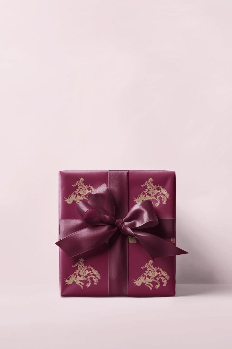 May include: A burgundy gift box with a matching ribbon and bow. The wrapping paper has a repeating gold illustration of a cowboy on a horse. The gift is set against a light pink backdrop. The box is suitable for various occasions.