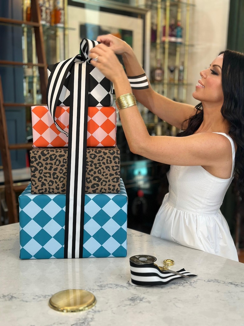 May include: A stack of wrapped gifts with various patterns, including a black and white diamond pattern, orange and white diamond pattern, leopard print, and blue diamond pattern. The gifts are tied with a black and white striped ribbon.