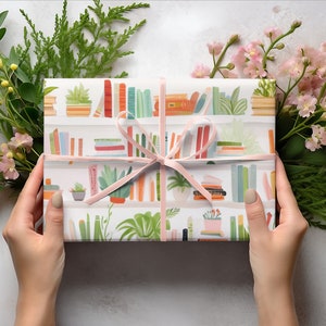 May include: A wrapped gift with a pink ribbon, featuring a bookshelf pattern with colorful books and potted plants. The gift is surrounded by green foliage and pink flowers, creating a decorative presentation.