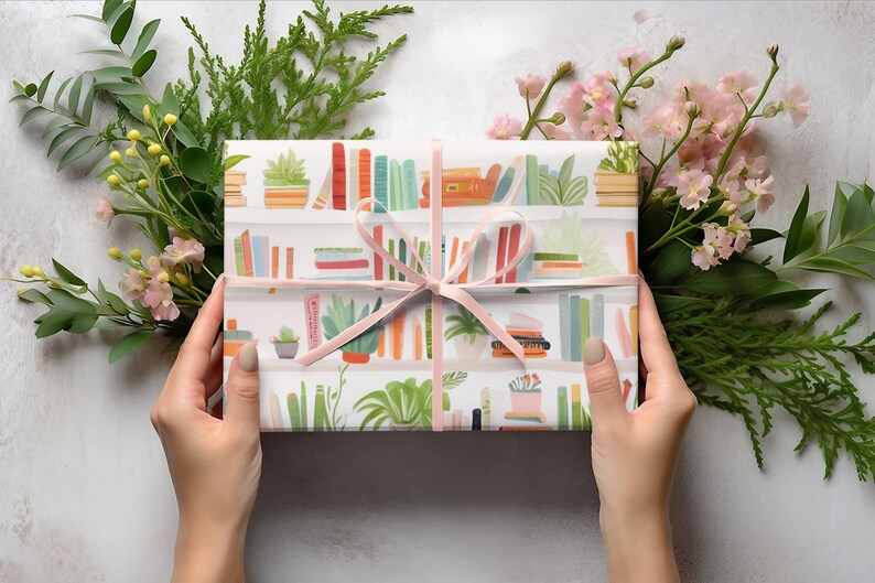 May include: A wrapped gift with a pink ribbon, featuring a bookshelf pattern with colorful books and potted plants. The gift is surrounded by green foliage and pink flowers, creating a decorative presentation.