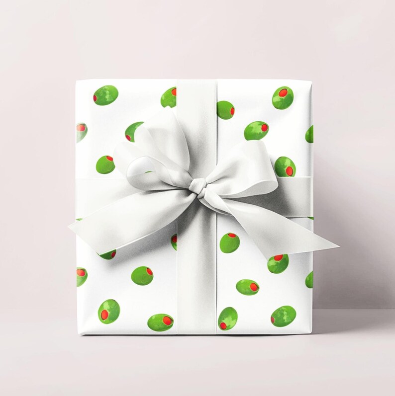 May include: A wrapped gift with white paper featuring a pattern of green olives with red centers. A large white satin ribbon is tied around the gift in a bow. The gift is presented on a light pink background.