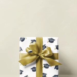 K&ouml;nnte beinhalten: Eine wei&szlig;e Geschenkbox mit goldener Schleife und einem Muster aus Abschlusskappen. Die Box ist in wei&szlig;es Papier mit einem sich wiederholenden Muster aus schwarzen Abschlusskappen mit goldenen Quasten verpackt.