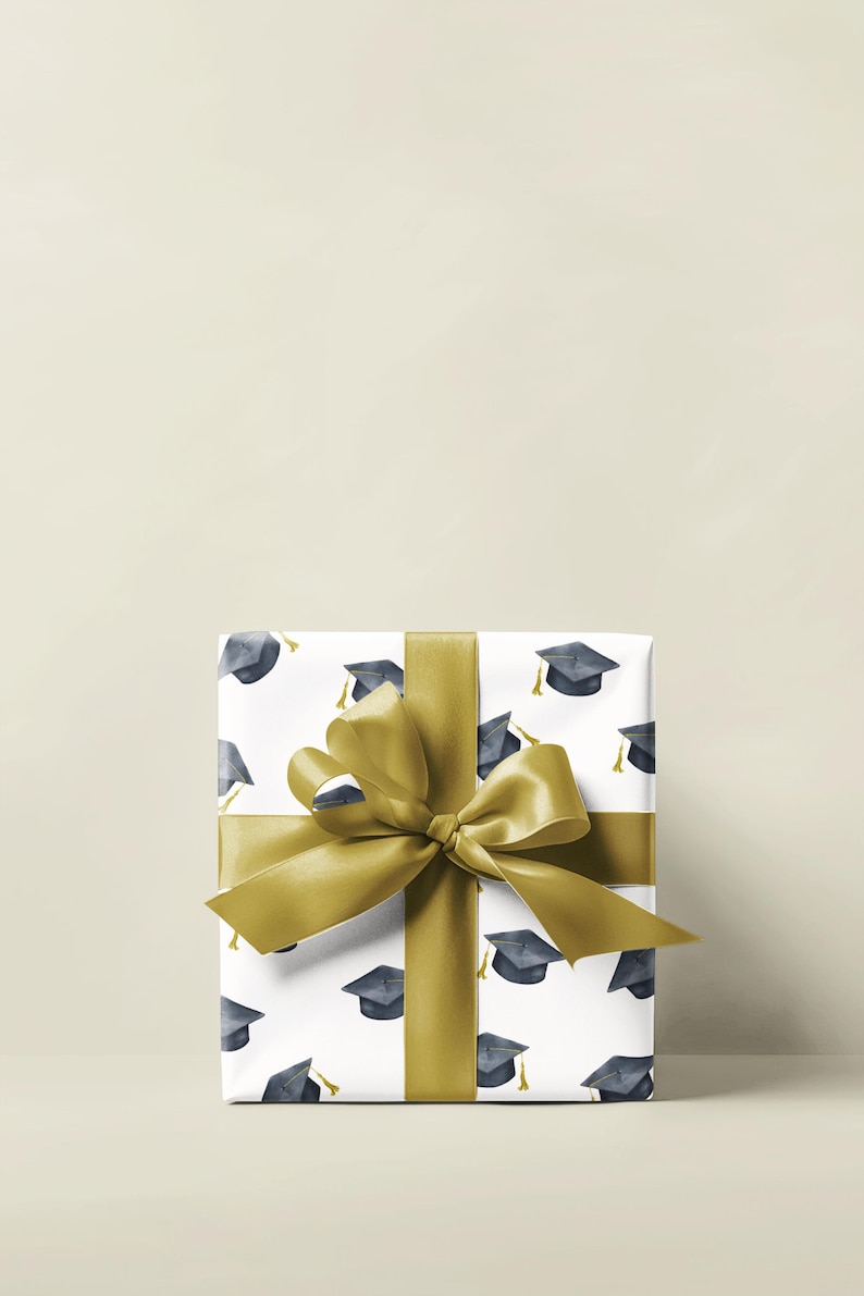 K&ouml;nnte beinhalten: Eine wei&szlig;e Geschenkbox mit goldener Schleife und einem Muster aus Abschlusskappen. Die Box ist in wei&szlig;es Papier mit einem sich wiederholenden Muster aus schwarzen Abschlusskappen mit goldenen Quasten verpackt.