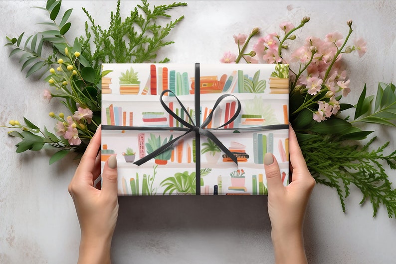 May include: A wrapped gift with a black ribbon, featuring a bookshelf pattern with colorful books and potted plants. The gift is surrounded by green foliage and pink flowers, creating a decorative presentation.