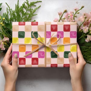 K&ouml;nnte beinhalten: Eine Geschenkbox, verpackt in rosa und braunes kariertes Papier mit bunten Quadraten. Das Papier hat einen Aquarell-Effekt und die Box ist mit einem braunen Band gebunden.