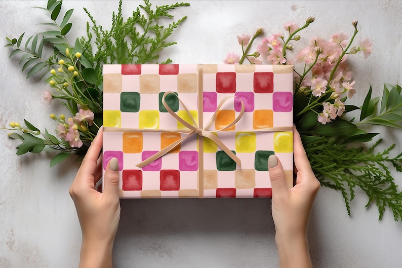 K&ouml;nnte beinhalten: Eine Geschenkbox, verpackt in rosa und braunes kariertes Papier mit bunten Quadraten. Das Papier hat einen Aquarell-Effekt und die Box ist mit einem braunen Band gebunden.