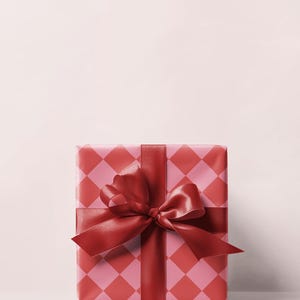 May include: A wrapped gift box with a red and pink diamond pattern. The box is tied with a wide, red satin ribbon and bow. The background is a solid, light pink color.
