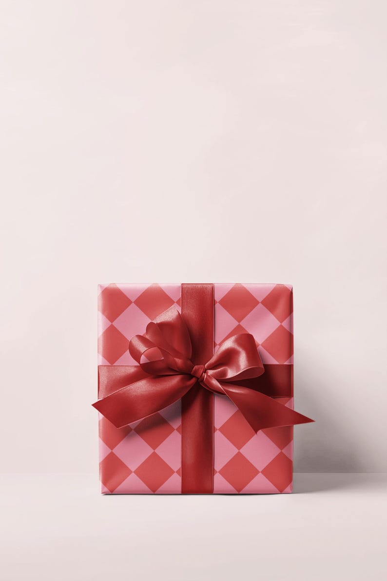 May include: A wrapped gift box with a red and pink diamond pattern. The box is tied with a wide, red satin ribbon and bow. The background is a solid, light pink color.